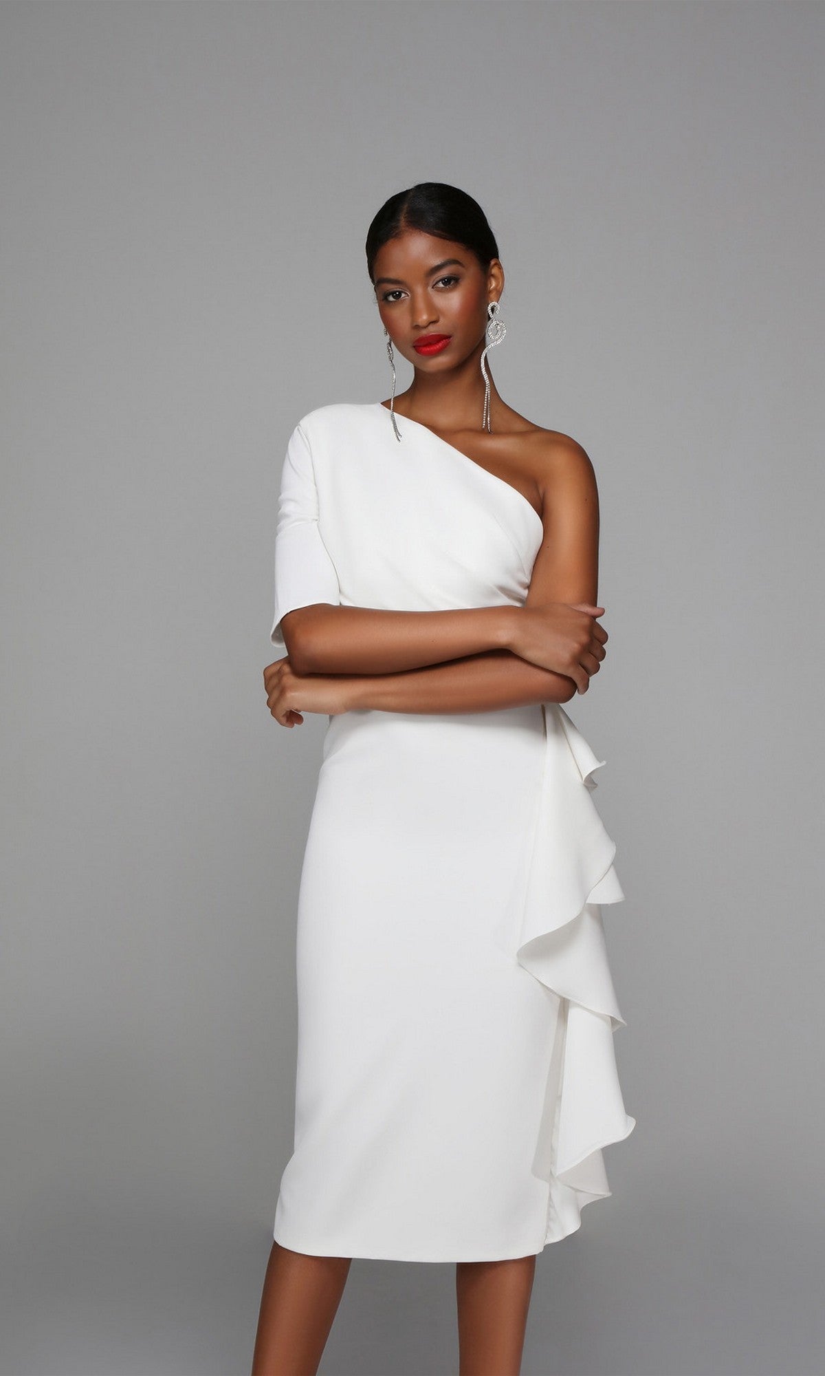 A woman poses with arms crossed, wearing the Knee Length Semi-Formal Dress: Alyce 70006, featuring a one-shoulder design and ruffle detail on the skirt, against a plain grey background.